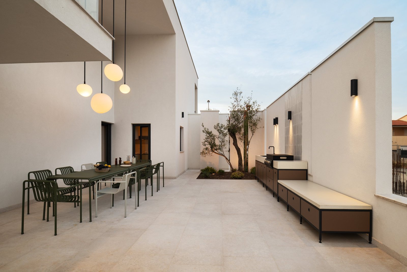 Arc House: terrace with a long dining table and olive tree at dusk